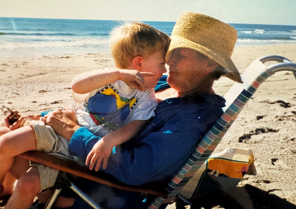 A young child playfully touching the nose of an older woman sitting on a beach chair by the ocean, both smiling, with the sun shining and a beach backdrop.